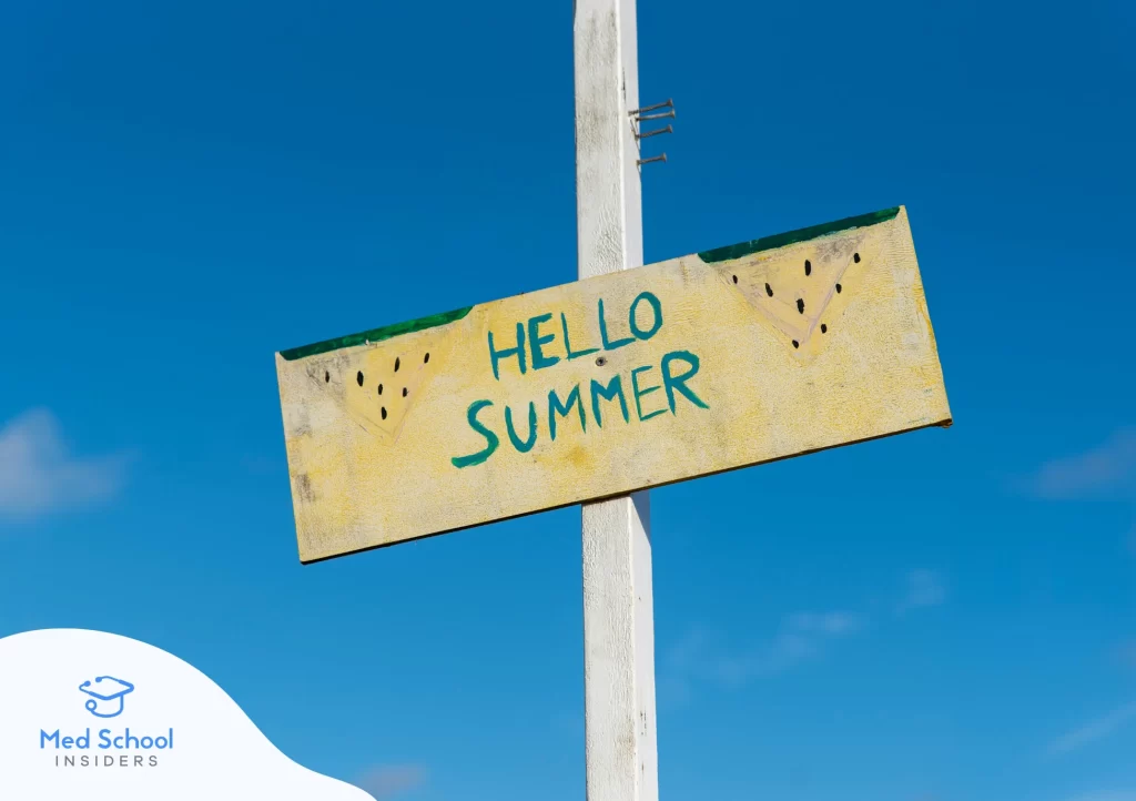 A weathered yellow wooden sign reading 'Hello Summer' with watermelon slice decorations, mounted on a white post against a bright blue sky. Med School Insiders logo in the bottom left corner.