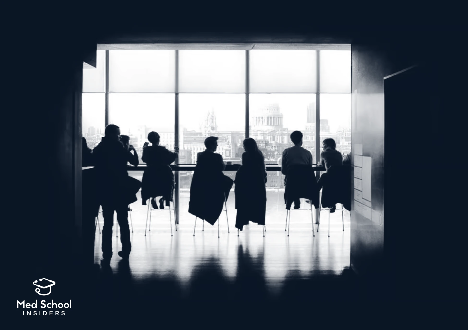 Silhouettes of a group of people seated and standing at a long table in front of large windows overlooking a city skyline, in a dark, moody black-and-white photo. Med School Insiders logo in the bottom left corner